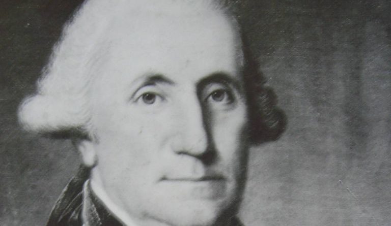 На данашњи дан: Црно-бели портрет мушкарца са белом, коврџавом косом, у капуту са високом крагном и кравати, који гледа директно у посматрача на обичној, тамној позадини.